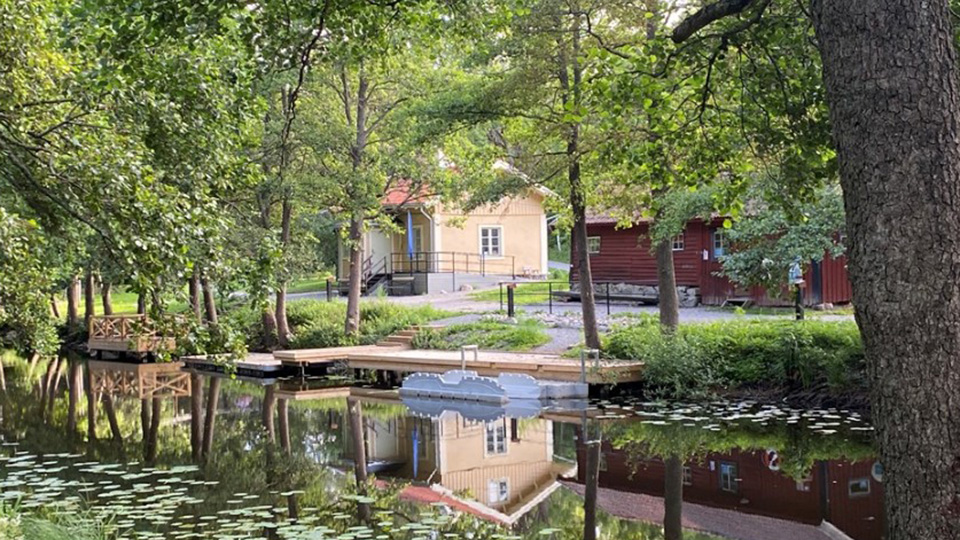 Ett trähus vid Åkers kanal. Det är sommar, tät grönska och huset speglas i vattnet. Det finns en brygga med ramp för kanot.