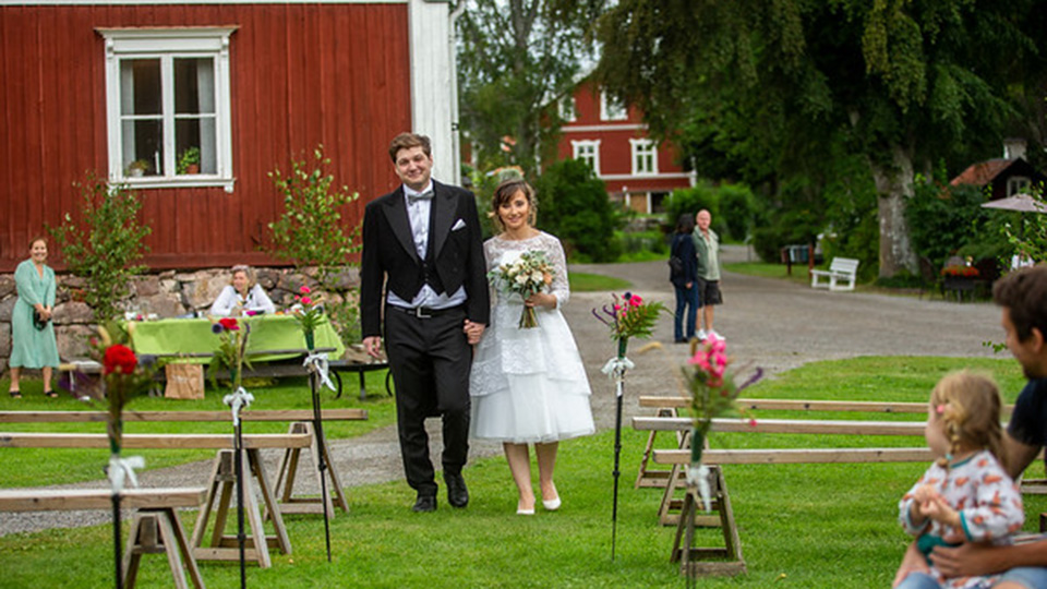 En brudgum i frack och en brud i vit klänning. Det är utomhus, sommargrönt och de går leende mot kameran.