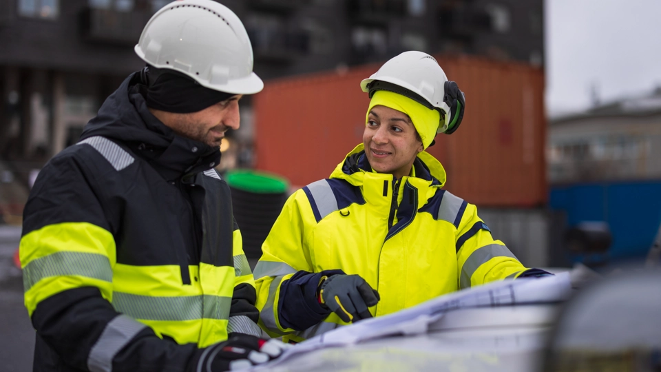 En kvinna och en man står i varselkläder och blickar ut på ett stort pappersark. På huvudet har båda personerna vita bygghjälmar och i bakgrunden kan man skymta containrar.