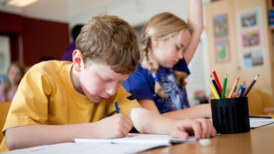 Två elever sitt vid sin skolbänk. En räcker upp handen, den andra skriver i sin skrivbok.