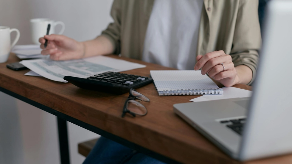 En person sitter vid ett skrivbord. På skrivbordet finns det papper, anteckningsblock, miniräknare, glasögon, dator och två kaffekoppar. Personen håller i en penna i sin högra hand.