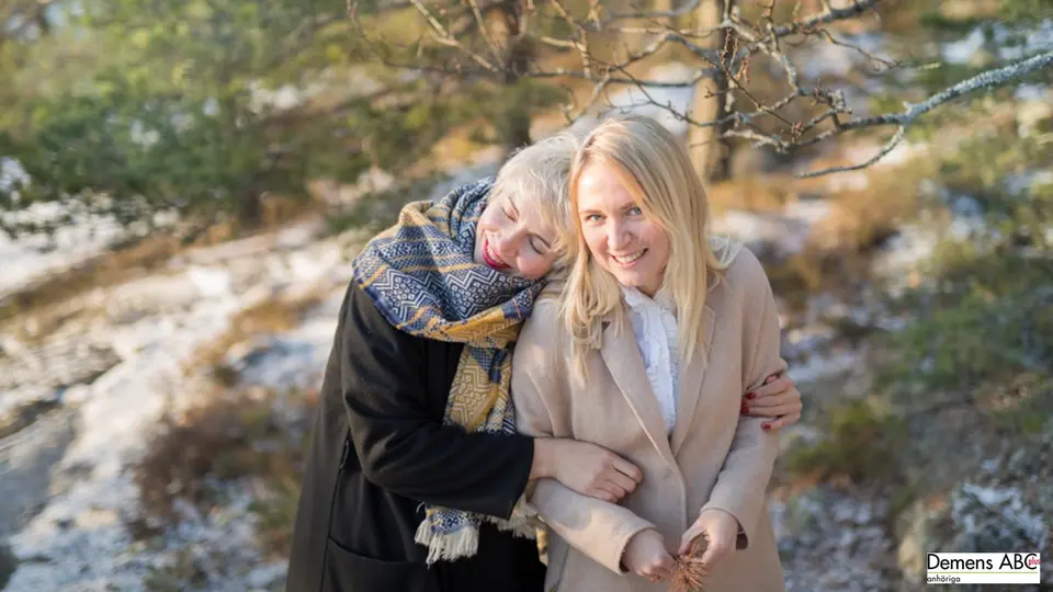 En kvinna sitter bredvid en blommande krukväxt. Bredvid henne, med huvudet lutat mot den äldre kvinnans axel sitter en yngre.