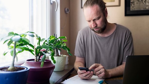 Man vid köksbord kollar mobilen framför datorn