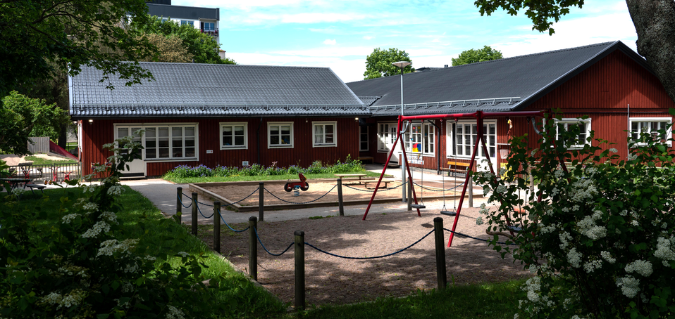 Färglådans förskola, röd träbyggnad med en sandlåda framför, buskar med vita blommor i förgrunden