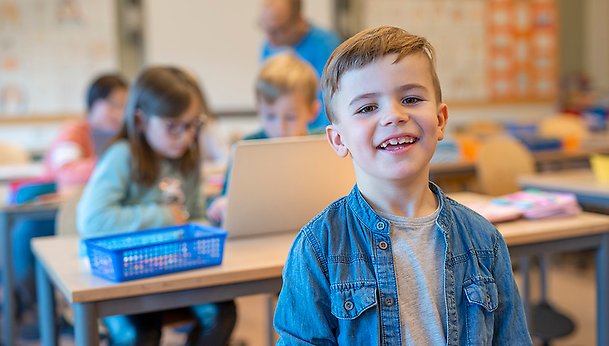 Barn sitter i ett klassrum och ler mot kameran, bär en blå tröja