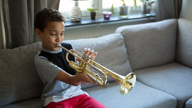 Barn med grå/svart tröja och röda byxor sitter i soffa och spelar trumpet