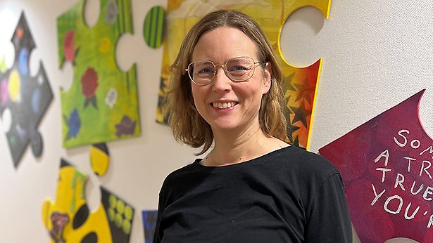 Porträtt på Anette Sundström som står mot en vit vägg med flerfärgade pusselbitar på.