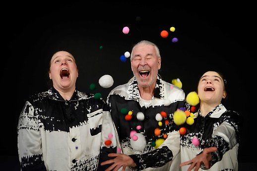 Tre personer gapskrattar medan det regnar ned små bollar över dem.