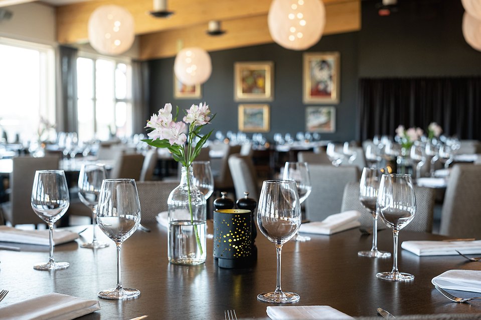 Flera dukade bord på en restaurang. Höga vinglas och snittblommor.