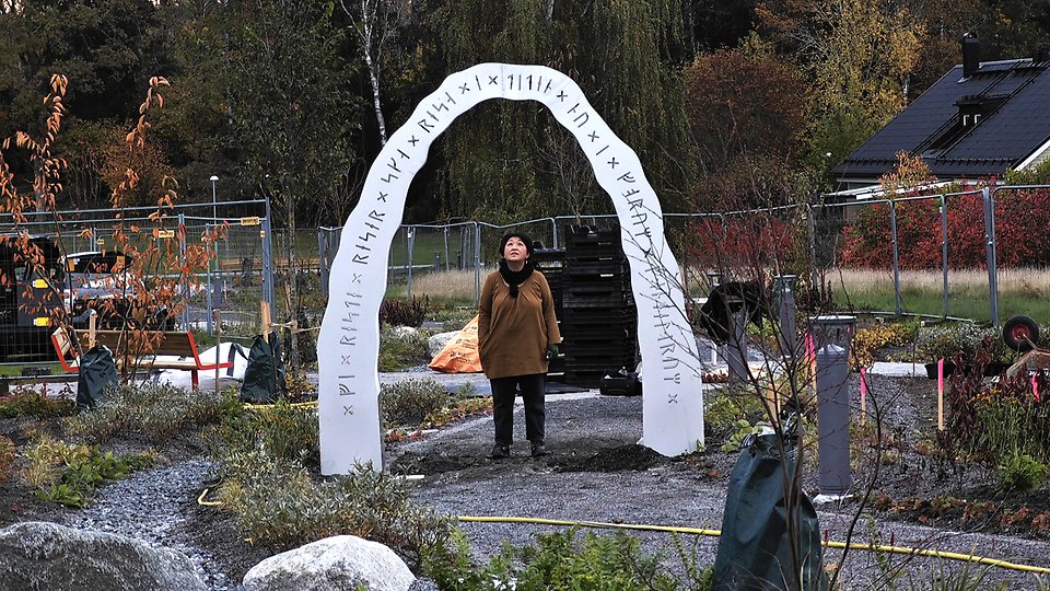 Bild på en bågformad installation, Tidsportalen, med runskrift ingraverad i Skånstaparken. Runskriften  lyder: ”Vi ska leka, leker, lekte, nu i Forum Vendelorum. Konstnären Guangjuan Zhang är med på fotot.