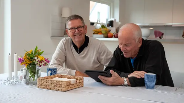 Två personer sitter vid ett bord. den ena ler in i kameran, den andra tittar ner i en läsplatta.