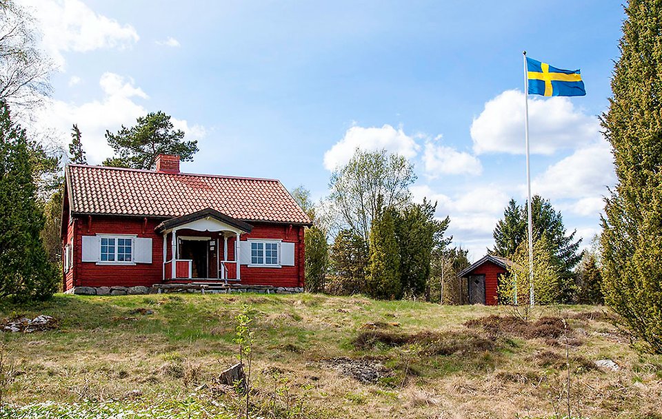 En röd trästuga med röda knutar. En svenska flagga är hissad bredvid.