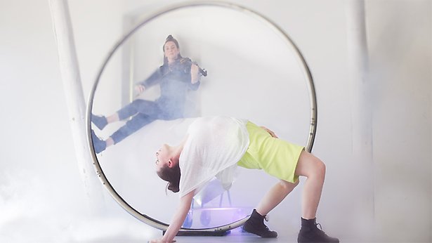 Två artister: en sitter i bakgrunden och spelar fiol. Framför henne utför en akrobat konster med en stor ring.