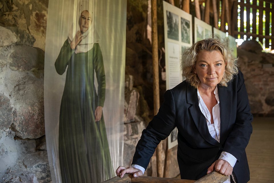Porträttbild av Elisabeth Ohlson står i Bisopstuna borgruin med utställningen synlig bakom sig