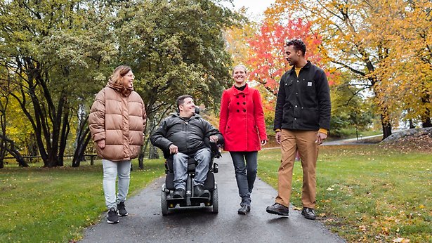 Fyra personer går i en park. En av dem är rullstolsburen.