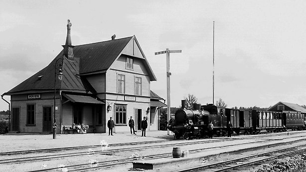 Ett svartvitt äldre foto som visar en stilig stationsbyggnad i trä. Det står Åkers-Berga på skylten. Framför står ett ånglok med vagnar.