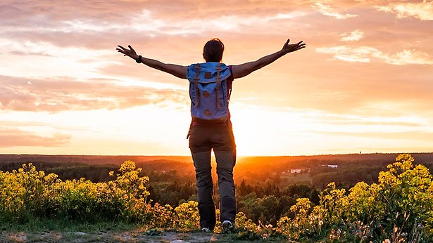En person står med ryggen mot kameran och med armarna utsträckta mot sidorna. Personen står på toppen av ett berg och tittar ut mot dalen, längst bort i bild syns en vacker solnedgång. 