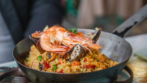 En man sitter på en restaurang med en paellapanna framför sig toppad med en stor räka. 