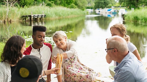 En grupp ungdomar och vuxna sitter på en picknickfilt med Åkers kanal i bakgrunden.