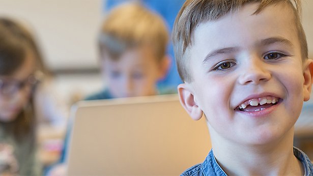 En glad pojke i ett klassrum tittar in i kameran. I bakgrunden syns två elever som jobbar tillsammans.