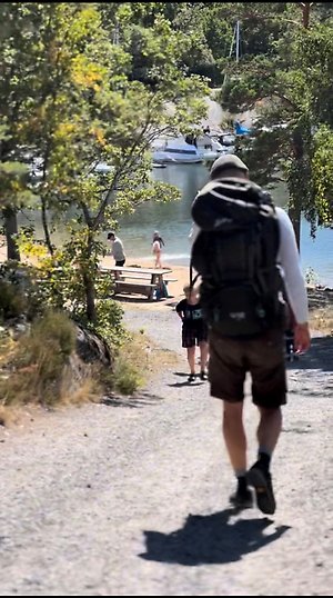 Man med ryggsäck vandrar på grusväg. En sandstrand och havsvik med segelbåtar syns i bakgrunden. Det är en solig sommardag.