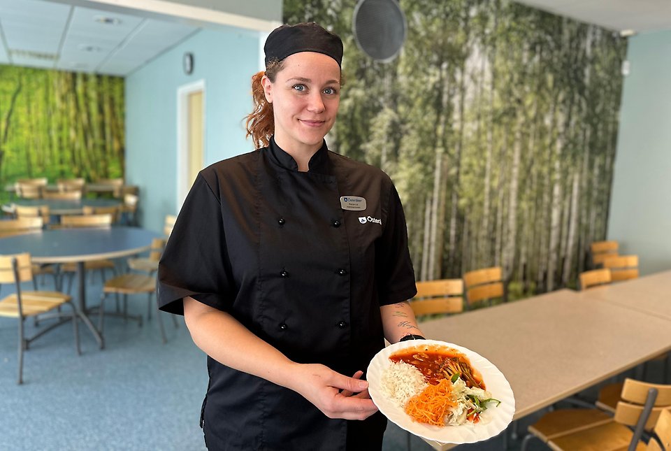 Bilden visar Rebecka Brolin i svarta kockkläder som står i matsalen och håller upp en tallrik med dagens lunch.