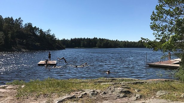 Klippor i förgrunden och en liten flytbrygga i sjön där barn leker och dyker. Det är en solig sommardag.