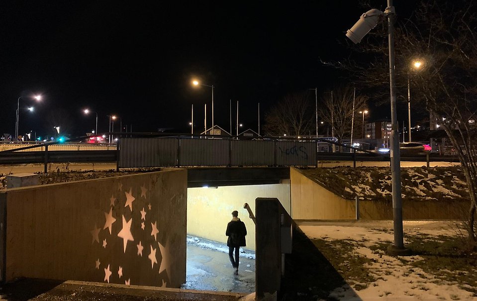 Bilden visar en person bakifrån som går in i tunneln under cirkulationsplatsen vid Cirkel-K (Roslagsvägen/väg 276, Norrgårdsvägen och Söralidsvägen). Det är mörkt och lite snö ligger på marken. Vid trappen ner till öppningen av tunneln syns en trygghetsstärkande insats - ljussättning med stjärnor för en tryggare upplevelse av platsen.