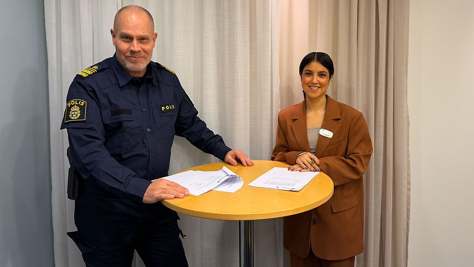 Två personer står vid ett runt bord och skriver under dokument, en polis i uniform och en kvinna i kavaj.