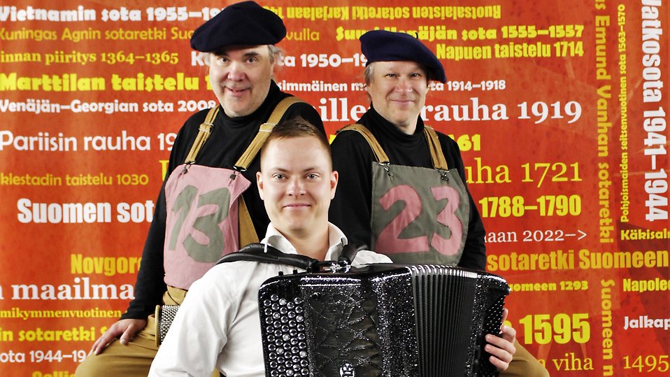 Två män med var sin basker står bakom en sittande man med dragspel. I bakgrunden en fond med olika ord på finska.