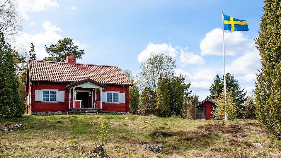 Rödmålad timmerstuga på en höjd med intilliggande bod och med svenska flaggan hissad i flaggstången