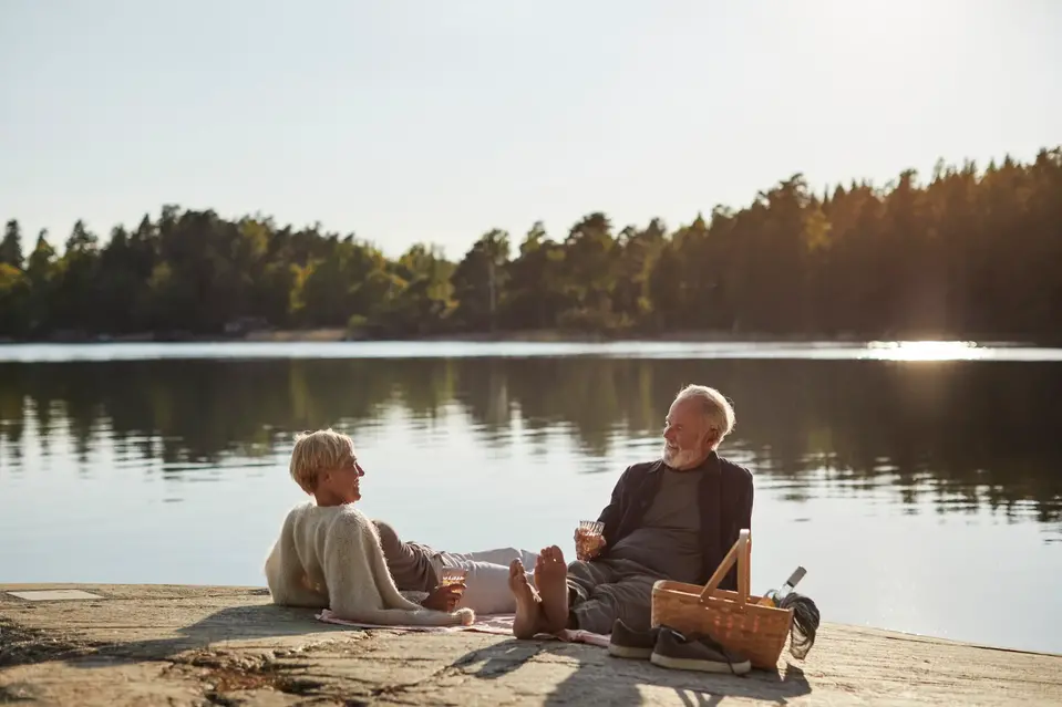 Äldre par sitter på en filt vid en sjö och dricker något tillsammans i kvällssolen. En picknickkorg står bredvid dem och skog syns på andra sidan vattnet.