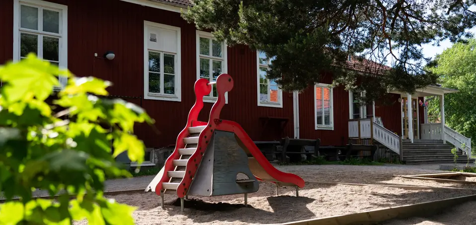 Bild på Roslags-Kulla förskola, röda enplans hus som sitter ihop i grönskande miljö