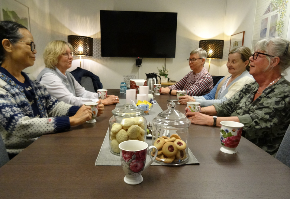 Fem personer sitter runt ett bord och samtalar i en anhöriggrupp. På bordet finns kaffekoppar och kakor i en lugn och hemtrevlig miljö.